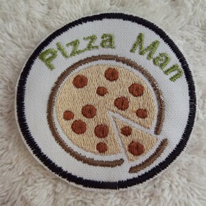 May include: A white embroidered patch with a black border. The patch features a pizza with brown pepperoni and a green inscription that reads "Pizza Man".