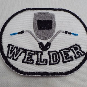 Può includere: Una toppa ovale bianca ricamata con un contorno nero. La toppa presenta un casco da saldatore grigio con una visiera nera e due torce da saldatura blu. La parola "WELDER" è ricamata in nero sotto il casco.