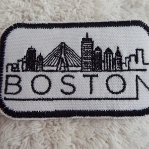 May include: White embroidered patch with a black outline. The patch features a skyline of Boston, Massachusetts, with the word "BOSTON" written below the skyline.