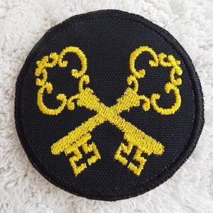 May include: A black circular patch with two gold embroidered keys crossed over each other. The keys have ornate designs.