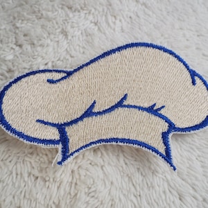 Può includere: Toppa ricamata a forma di cappello da chef. La toppa presenta un cappello color crema con un contorno blu. Il cappello ha la forma classica di un cappello da chef, con una parte superiore arrotondata e un bordo. Il contorno blu aggiunge colore.
