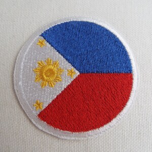 May include: A circular embroidered patch featuring the flag of the Philippines. The flag is white with a blue triangle at the hoist and a red triangle at the fly. A yellow sun with eight rays and three stars is in the center of the white field.