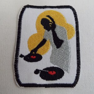 May include: An embroidered iron-on patch featuring a black silhouette of a person with headphones on, spinning two black vinyl records on a turntable. The background is a gold and silver design.