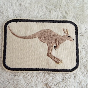 Può includere: Un canguro ricamato marrone su una toppa di stoffa bianca con un bordo nero.