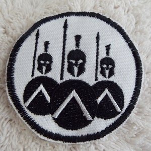 Puede incluir: Un parche bordado blanco con tres cascos y escudos espartanos negros. Los cascos tienen plumas blancas y los escudos tienen triángulos blancos.