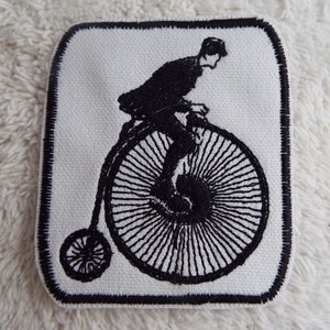 May include: Black and white embroidered patch of a person riding a penny-farthing bicycle.