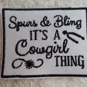 May include: White embroidered patch with black stitching. The text reads "Spurs & Bling It's a Cowgirl Thing".