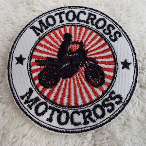 May include: A white, round embroidered patch with a black and red motocross design. The patch features a silhouette of a motocross rider on a dirt bike, with a red and white sunburst design behind it. The words "MOTOCROSS" are embroidered in black around the edge of the patch.
