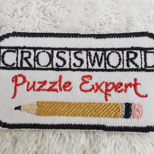 Könnte beinhalten: Weißer gestickter Aufnäher mit schwarzen Nähten. Der Aufnäher zeigt den Text "CROSSWORD Puzzle Expert" in schwarzen und roten Buchstaben. Unter dem Text ist ein gelber Bleistift gestickt.