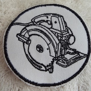 May include: A white circular patch with a black embroidered image of a circular saw. The saw is outlined in black thread and has a detailed design.