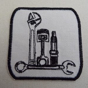 May include: Embroidered patch with mechanic's tools: a spanner, piston, and spark plug. Black design on a white background, with a black border. Suitable for automotive enthusiasts or mechanics.