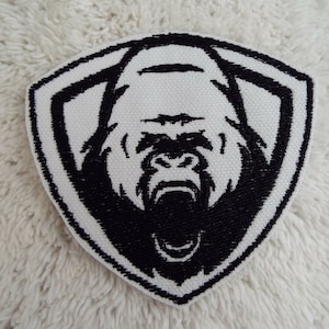 Puede incluir: Parche bordado en blanco y negro de la cara de un gorila con la boca abierta, sobre una forma de escudo blanco.