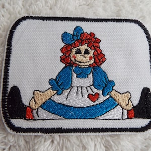 May include: Embroidered patch featuring a red-haired rag doll with a blue dress and a red heart on her chest. The doll is sitting with her legs crossed and has black shoes.