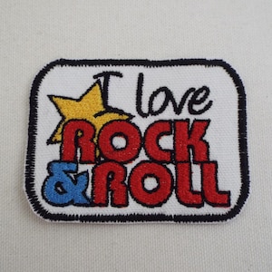 Puede incluir: Parche bordado con el texto "I love Rock & Roll". El parche presenta una estrella amarilla, letras rojas y azules y un borde negro. El parche es rectangular con esquinas redondeadas.