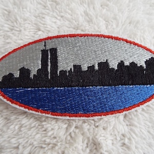 May include: An embroidered patch with a red, white, and blue border. The patch features a black silhouette of the New York City skyline, including the Twin Towers.