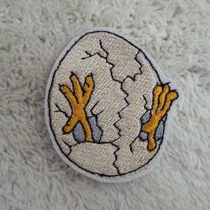 May include: A white embroidered patch of a chick hatching from an egg. The chick's yellow feet are visible as it breaks out of the egg.