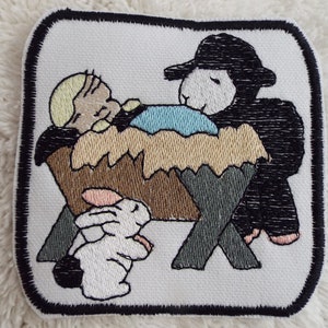 May include: Embroidered patch depicting a nativity scene with a baby in a manger, a sheep, and two white rabbits. The patch is white with black stitching around the edges.