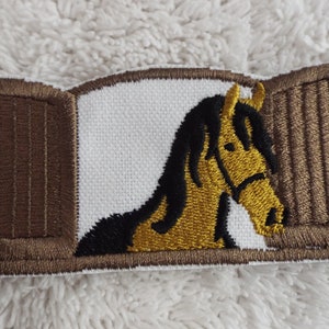 Könnte beinhalten: Gestickter Aufnäher mit einem braunen Pferd mit schwarzer Mähne und Schweif, das aus einer braunen Holztür eines Stalls schaut.