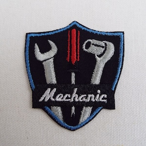 以下が含まれることがあります： 黒地に青と白の縁取りのある盾の形をした刺繍パッチ。盾の中にはレンチ、ドライバー、ソケットレンチの3つの工具が入っています。盾の下部には黒いバナーに白い文字で「メカニック」と書かれています。