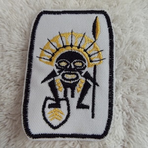 Puede incluir: Un parche bordado blanco con un diseño tribal negro y dorado de una figura sosteniendo una lanza. La figura lleva un tocado con un diseño de sol.