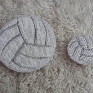May include: Two white embroidered volleyball patches, one larger than the other, on a white fuzzy surface.