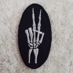 May include: A black oval-shaped patch with a white embroidered skeleton hand making the peace sign.