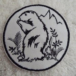 May include: Black and white embroidered patch of a prairie dog sitting up with its paws in front of it. The prairie dog is surrounded by grass and a mountain in the background.