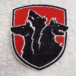 Puede incluir: Un parche bordado rojo y negro con un diseño de lobo de tres cabezas. Las cabezas del lobo miran en diferentes direcciones, y el parche tiene forma de escudo.