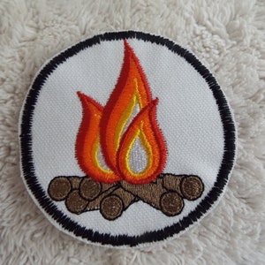 May include: Embroidered patch featuring a campfire design. The patch has a white background with a black border. The flames are red, orange, and yellow, and the logs are brown. This patch is suitable for jackets, bags, or other fabric items.