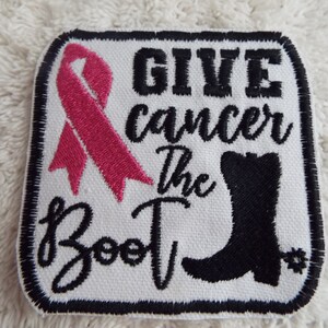 Puede incluir: Parche de tela blanca con costuras negras y una cinta rosa. El texto dice "GIVE cancer the BOOT" con un gráfico de bota negra.