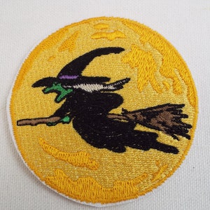 May include: A yellow embroidered patch with a black witch flying on a broomstick in front of a full moon. The witch is wearing a black hat with a purple band and a green face.