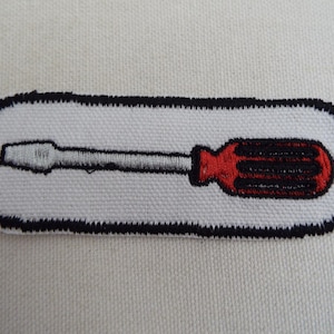 May include: A white embroidered patch with a black outline depicting a red and black screwdriver.