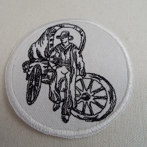 May include: Black and white embroidered patch depicting a cowboy riding a wagon with a large wheel. The patch is circular and has a white background.