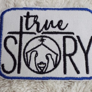 May include: White embroidered patch with blue edging. The patch features a black and white illustration of the nativity scene with the text "true story" above it.