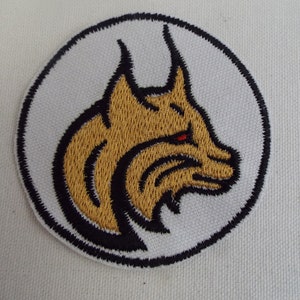 May include: A white embroidered patch with a black outline, featuring a gold-colored lynx head with red eyes. The lynx is facing right and has a detailed, stylized design.
