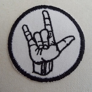 May include: Embroidered patch featuring a black outline of a hand making the "I love you" sign within a white circle. The circle is bordered by a black stitched edge. The patch is likely for clothing or accessories.