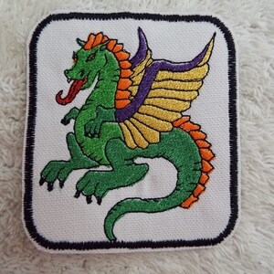 May include: Embroidered iron-on patch featuring a green dragon with orange and yellow wings, purple accents, and a red tongue. The dragon is on a white background with a black border.