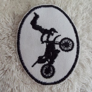 Pode incluir: Remendo oval com um desenho bordado a preto de uma pessoa a fazer uma acrobacia numa mota. O remendo tem um fundo branco e uma borda cosida a preto. A mota está no ar, com os braços e as pernas do piloto esticados.