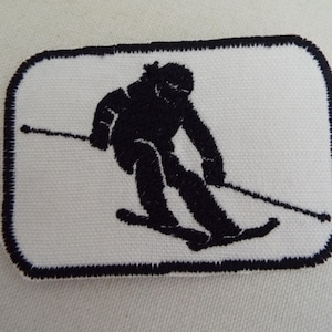 Peut inclure: Patch brodé noir et blanc d'un skieur sur fond blanc. Le skieur est en position de descente avec des skis et des bâtons de ski.