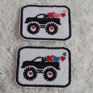 May include: Two embroidered iron-on patches featuring a black monster truck carrying a load of hearts. The top patch has pink and red hearts, while the bottom patch has red and blue hearts.