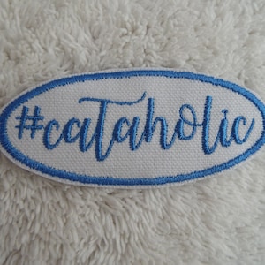 Puede incluir: Parche ovalado blanco con borde azul y el texto "#catholic" bordado en azul.