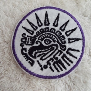 May include: A white embroidered patch with a black design of a stylized face with a sunburst around it. The patch has a purple border.