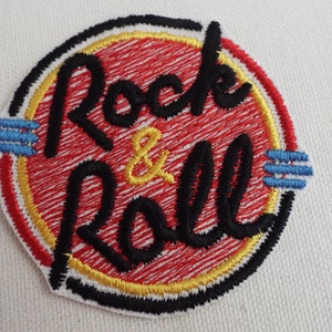 Puede incluir: Un parche bordado rojo, amarillo y negro con las palabras "Rock & Roll" en un círculo.