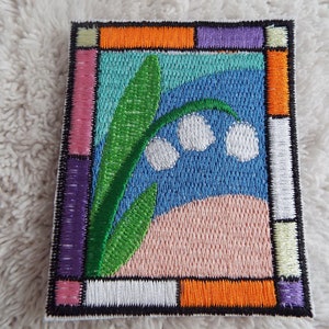 Könnte beinhalten: Gestickter Aufnäher mit einem Buntglasdesign, das eine weiße Maiglöckchenblume mit grünen Blättern auf blauem Hintergrund zeigt. Der Rahmen besteht aus bunten Quadraten in Rosa, Lila, Orange und Weiß.