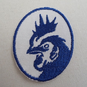 May include: Embroidered patch featuring a blue rooster design within a white oval. The rooster is depicted in profile, with detailed feathers and a prominent comb. The patch is likely for clothing or accessories.