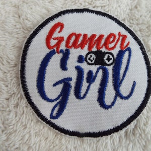 May include: A white round patch with a black border. The words "Gamer Girl" are embroidered in blue and red. A black and white video game controller is embroidered in the centre of the patch.