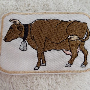 May include: Embroidered patch of a brown cow with a bell around its neck. The cow is standing on a white background with a beige border.