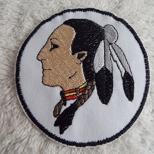 Puede incluir: Un parche bordado blanco con un contorno negro que representa el perfil de un hombre nativo americano con la cara marrón, el pelo negro y un tocado de plumas blancas y negras.