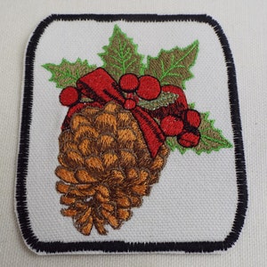 May include: Embroidered iron-on patch featuring a brown pine cone with a red bow and green holly leaves.