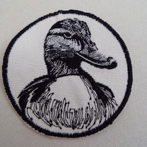 Puede incluir: Parche bordado con una ilustración en blanco y negro de la cabeza de un pato dentro de un marco circular. El parche está sobre una tela blanca, con un borde cosido negro. El diseño muestra la cabeza y la parte superior del cuerpo del pato.